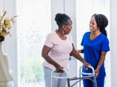istockphoto-1297322863-612×612-1 A healthcare worker visiting a senior African-American woman at home, helping her use a mobility walker. The patient is in her 70s and the nurse is a mature woman in her 40s.