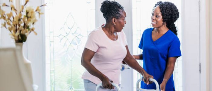 istockphoto-1297322863-612×612-1 A healthcare worker visiting a senior African-American woman at home, helping her use a mobility walker. The patient is in her 70s and the nurse is a mature woman in her 40s.